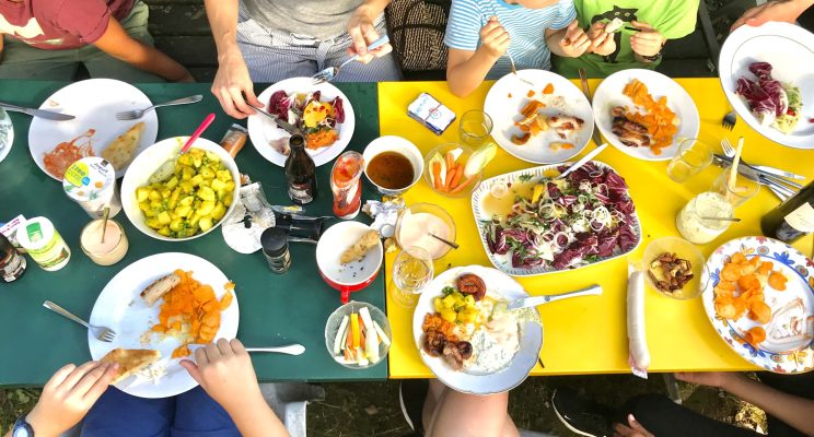Ein Buntes Foto einer gelben und grünen Tischplatte zu einem langen Tisch zusammengestellt auf dem viele Teller mit Erwachsenen und Kindern die dran sitzen und zusammen essen, von oben otografiert, man sieht keine Gesichter